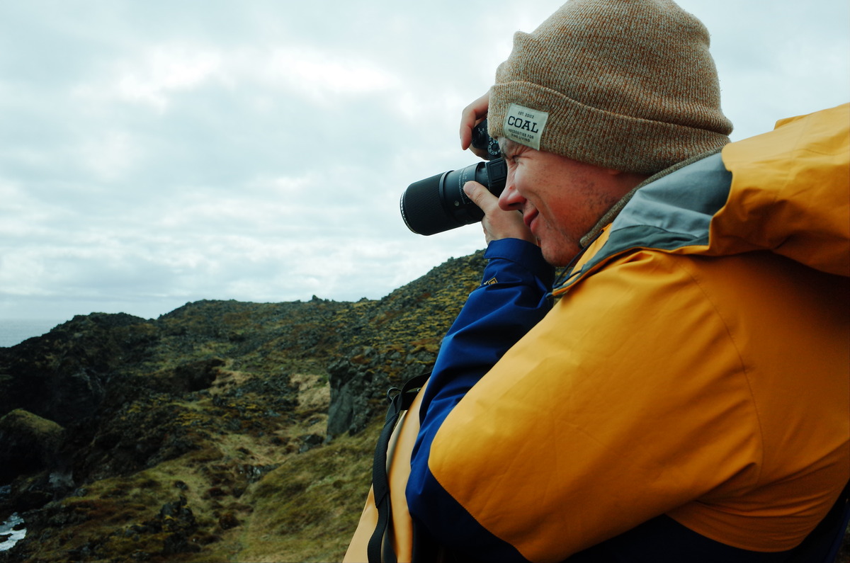 Person in a yellow and blue jacket with a brown beanie photographing a rocky landscape.