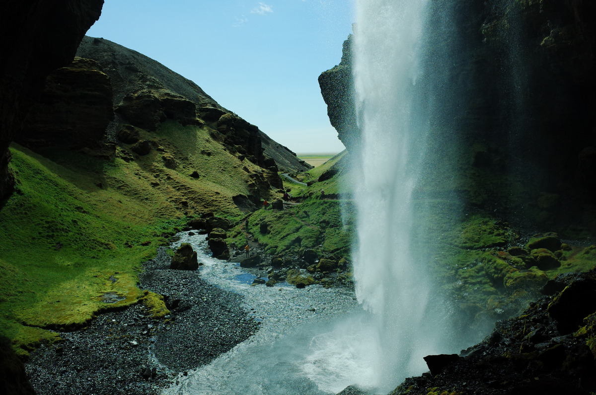 Upward angle of Kvernufoss as water plunges into a rocky stream surrounded by lush green hills.