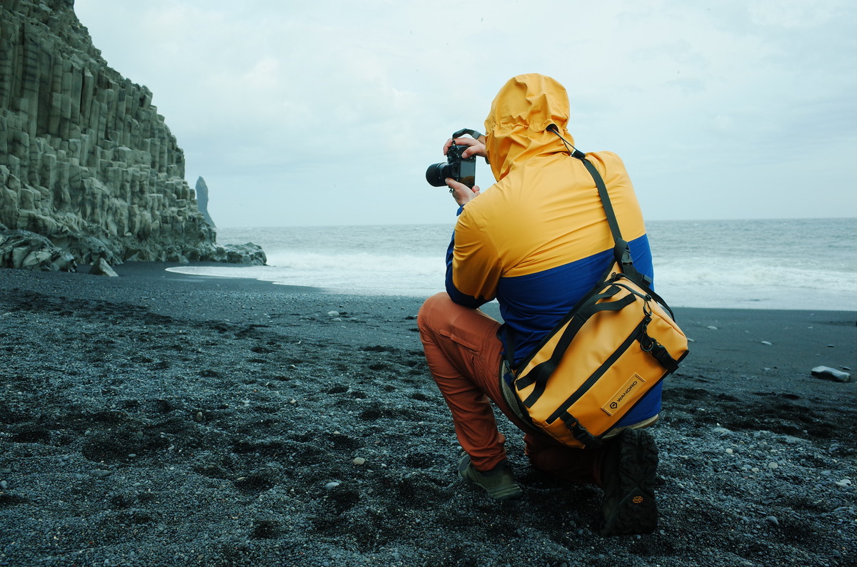 Photographer kneeling on the black sand beach of Reynisfjara, Iceland, with a camera raised to take a picture.