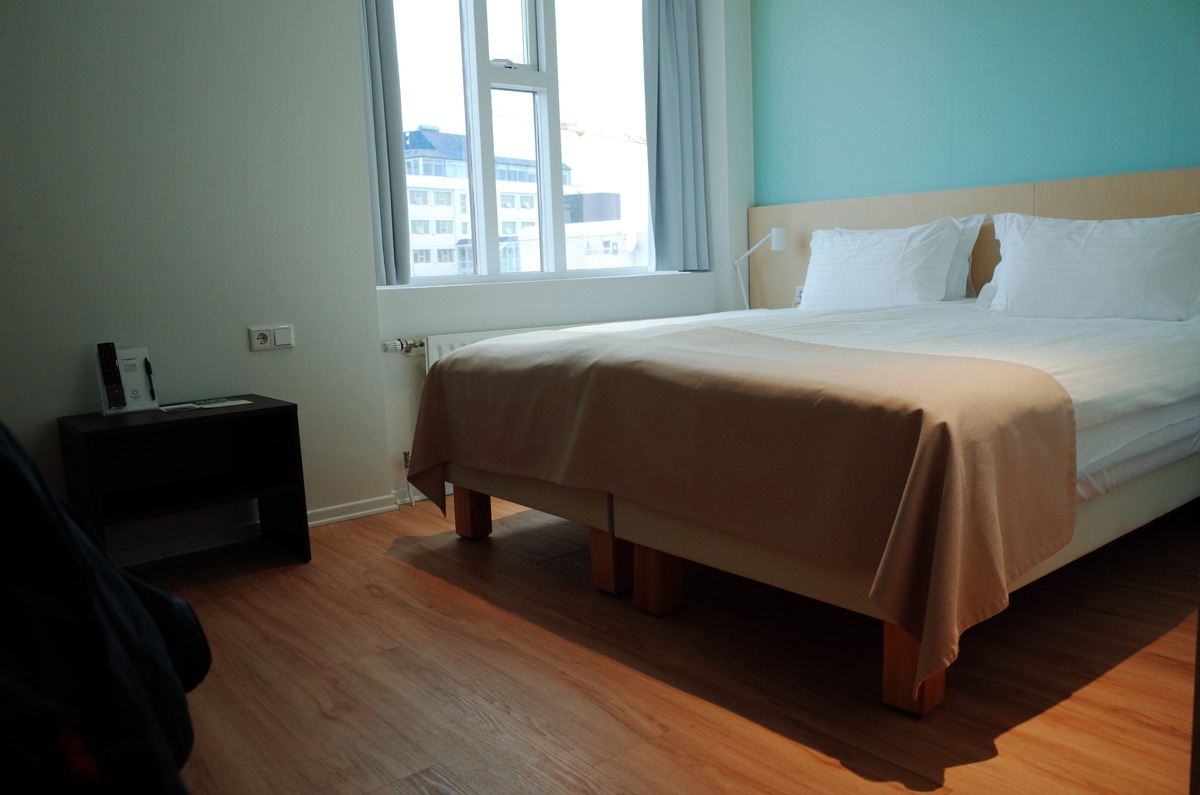 A bed next to a double pane window with a city view.