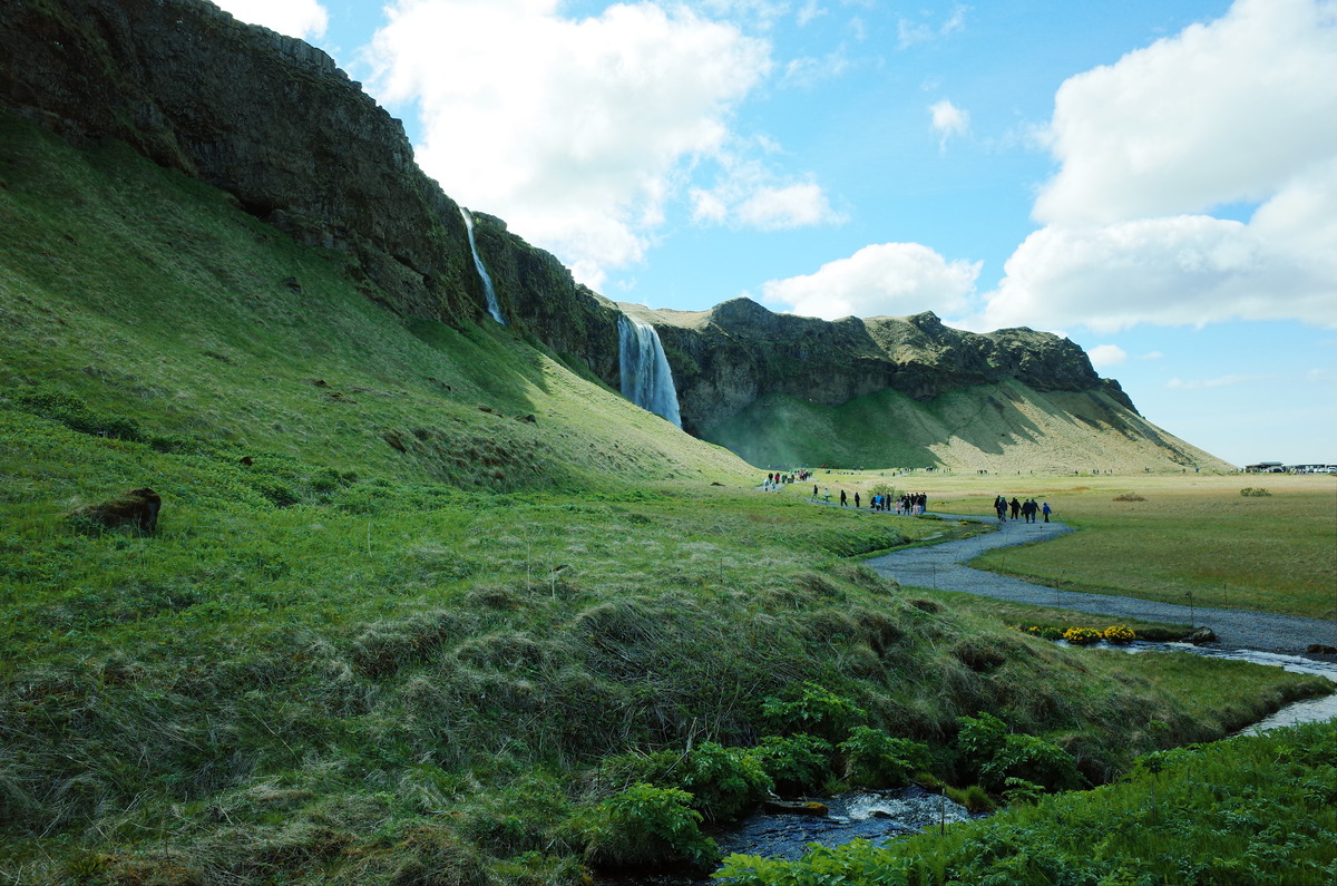 People walking on a gravel foot path next to the Seljalandsfoss waterfall.